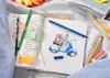 A flat lay of art supplies including pens, pencils, a sketchbook with a drawing of a scooter, and a notebook with writing. Produktbild-Vorschau