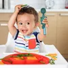 A baby sitting in a high chair, holding a dinosaur-shaped spoon and a piece of green vegetable on its head. There is a red plate with food in front of the baby. Produktbild-Vorschau