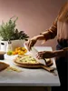A person is cutting a pizza on a wooden board on a marble countertop, surrounded by ingredients and herbs. Produktbild-Vorschau