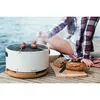 A person is sitting by the water, grilling food on a portable grill. Produktbild-Vorschau