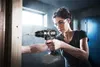 A woman is using a cordless drill to screw into a wooden plank in what appears to be a workshop or construction site. Produktbild-Vorschau