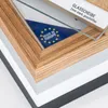 Close-up view of a picture frame corner, showing layers of wood, white, and gray material, with a glass pane and a 'Made in Europe' sticker. Produktbild-Vorschau