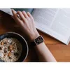 A person's wrist wearing a rose gold Apple Watch is shown next to a bowl of cereal and an open book. Produktbild-Vorschau