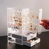 A hand is shown reaching for a clear acrylic jewelry organizer box filled with various necklaces and earrings. The organizer has two drawers at the bottom. Voir les images du produit