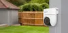 A white security camera is mounted on a gray pole in a backyard setting, with a wooden fence and greenery in the background. Produktbild-Vorschau