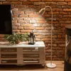 A floor lamp illuminates a living room area with a brick wall background, a TV stand made of pallets, and a plant. Anteprima dell’immagine prodotto