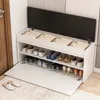 A white shoe cabinet with a black lid, filled with shoes and some folded items, is shown in a hallway next to a door. Podgląd zdjęcia produktu