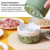 A hand is holding the top part of a green electric mini chopper over a clear container filled with chopped green ingredients. Plates with minced meat and other chopped food are visible in the background. Produktbild-Vorschau