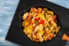 A close-up overhead view of a pan filled with mixed vegetables, placed on a black mat on a blue wooden surface. Náhľad obrázku produktu