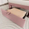A close-up view of a pink upholstered bed frame with a storage basket containing beige bedding underneath. Produktbild-Vorschau