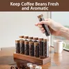 A hand holding a coffee bean storage tube above a wooden stand with multiple tubes filled with coffee beans. Produktbild-Vorschau