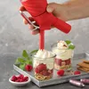 A hand holding a red whipped cream dispenser, dispensing whipped cream into two glasses filled with raspberries and cereal. Náhled obrázku produktu
