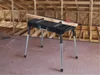 A black folding workbench with silver legs is set up in a room with unfinished wooden walls and ceiling, suggesting a construction or workshop environment. A piece of wood is clamped to the workbench. Náhľad obrázku produktu