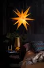 A star-shaped lamp hangs above a bedside table with a lamp, candles, and a pillow on the bed. Produktbild-Vorschau