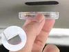 A hand is holding a small, clear, circular electronic device with internal components visible. A close-up inset shows the adhesive backing of the device, with a red protective layer being peeled off a gray foam pad. The device is being held up to a car's h Produktbild-Vorschau