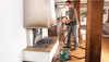A man is using a Bosch paint sprayer in a room with wooden beams and a fireplace. Produktbild-Vorschau