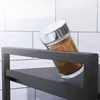 A spice jar with brown contents and a silver lid is tilted on a black shelf against a white tiled background. Náhľad obrázku produktu