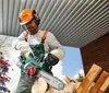 A man is using a chainsaw to cut a log outdoors, under a covered structure. Produktbild-Vorschau