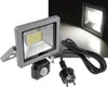 A floodlight with a motion sensor and a power plug is shown against a dark background, with a second floodlight illuminated in the background. Produktbild-Vorschau