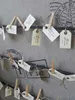 A close-up view of small wooden clothespins holding tags on a textured fabric background. Náhled obrázku produktu