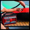 A person is using a red and black Einhell hybrid compressor to inflate a large red and black inflatable object, possibly a pool float or air mattress, on a wooden deck next to a turquoise pool. Voir les images du produit