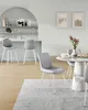 A modern dining and bar area with light blue walls, a white arched shelf, and light wood flooring. The scene features two grey bar stools at a white counter and a round white dining table with grey chairs, set with books and a vase of flowers. Náhled obrázku produktu