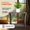 A small potted plant with orange fruits sits on a woven side table in front of a large window with a garden view. Produktbild-Vorschau