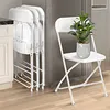 A set of white folding chairs are stacked in a kitchen or utility area next to a refrigerator. One chair is open and has a small potted plant on its seat. Podgląd zdjęcia produktu