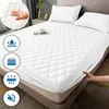 A white quilted mattress protector is shown on a bed, with pillows and a bedside table in the background. There are also icons on the left side of the image highlighting product features. Produktbild-Vorschau