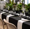 A beautifully set dining table with black tablecloth, white runner, black napkins with gold rings, and various plants in black pots. Produktbild-Vorschau