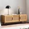 A wooden TV cabinet with cane-like doors and black legs, placed in a room setting with a lamp and a vase. Produktbild-Vorschau