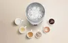 A white electric thermal vacuum hot water cup sits in the center, surrounded by seven small cups of various beverages. Some cups contain coffee or tea with toppings, while others appear to be plain water or light-colored beverages. Produktbild-Vorschau