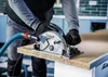A person wearing black gloves is using a Bosch circular saw to cut a piece of wood. A blue vacuum hose is attached to the saw. Voir les images du produit