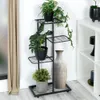 A black metal plant stand with four tiers, holding several potted plants and a large vase, placed indoors next to a beige curtain and a white shelving unit. Náhled obrázku produktu