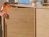 A person's hand is shown placing an orange book into a wooden cabinet. Náhľad obrázku produktu
