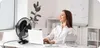 A woman sitting at an office desk with a laptop and a fan. Anteprima dell’immagine prodotto