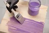 A hand wearing a glove is painting a wooden surface with purple paint. Voir les images du produit