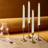 Four LED taper candles in brass candle holders are arranged on a marble surface, with a glass carafe and a small glass sphere to the left. The background is a textured brown fabric. Produktbild-Vorschau