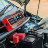 A person wearing a glove is using a car battery charger connected to a car battery under the hood of a car. Produktbild-Vorschau