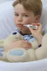 A young boy is lying in bed, holding a teddy bear and taking his temperature with a digital thermometer. Podgląd zdjęcia produktu
