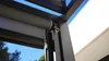 Close-up of a roller blind mechanism attached to a dark metal frame, with a blue sky and white building in the background. Produktbild-Vorschau