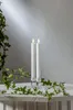 Two white LED taper candles with flickering flames stand in clear glass candle holders on a textured surface, with green ivy vines draped around them. Podgląd zdjęcia produktu