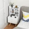 A white bedside table with drawers and shelves is placed next to a bed with a gray headboard and a colorful pillow. Podgląd zdjęcia produktu