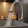 A kitchen sink with a flexible gray faucet spraying water into the sink. Jars of nuts and wooden bowls are visible in the background. Produktbild-Vorschau