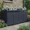 A dark gray, multi-unit trash bin enclosure with a planter on top, situated against a stone wall next to a building, with flowers and greenery surrounding it. Produktbild-Vorschau