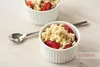 Two white ceramic ramekins filled with strawberry crumble are shown on a light-colored wooden surface, with a spoon to the left. Anteprima dell’immagine prodotto