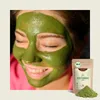 A woman with a green face mask applied, smiling with her eyes closed, next to a pouch of green powder. Produktbild-Vorschau