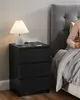 A person is sitting in bed next to a black nightstand with a lamp and a phone on it. Náhled obrázku produktu