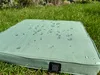 A green heated seat cushion with water droplets on it, resting on grass. Produktbild-Vorschau