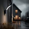 A modern outdoor wall lamp illuminates a pathway and house exterior at dusk. Produktbild-Vorschau